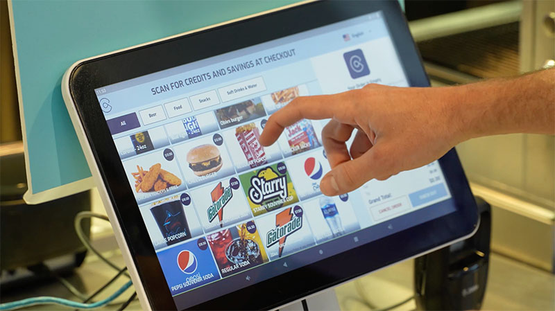 A hand selects a concession item for a customer on a touch screen kiosk.