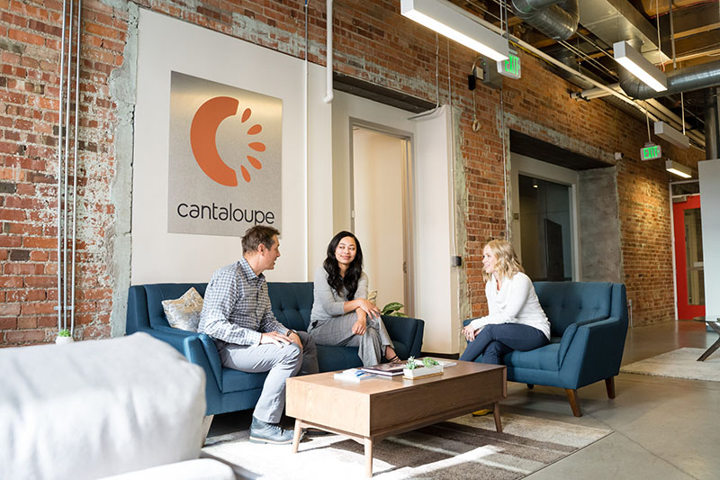 Three employees sit and chat in the Cantaloupe office lobby near a large Cantaloupe logo on the wall.