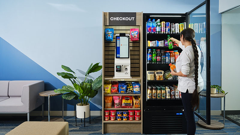 A woman selects items from a cool in a small micro market with a Go Plus100b kiosk.